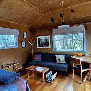 This cozy wooden cabin features a comfy sofa, small dining table, large windows, bird-themed decor, artwork, and natural light, creating a charming, inviting, and rustic atmosphere.