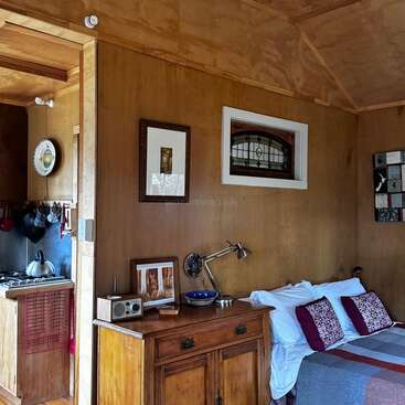 Um quarto aconchegante de madeira apresenta uma cama com travesseiros estampados, uma cômoda vintage com uma lâmpada, área de cozinha, arte na parede, vitral e decoração caseira.