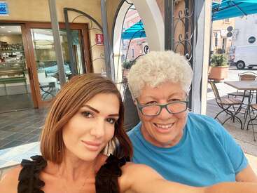 Zwei Frauen lächeln für ein Selfie in einem Straßencafé. Die Umgebung ist hell, entspannt und einladend, mit blauen Sonnenschirmen, Tischen und charmanten Straßenansichten.