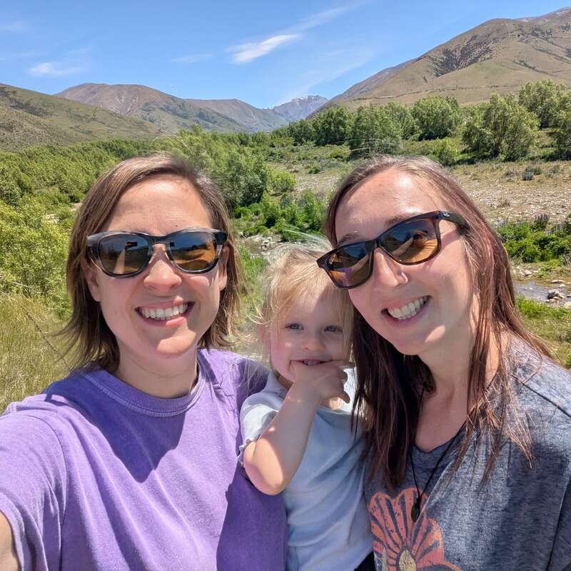Three people, two adults and a child, smile outdoors. They wear sunglasses, surrounded by green trees, mountains, and a blue sky under bright sunlight.
