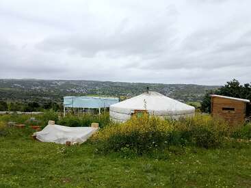 The image depicts a serene rural landscape featuring a white yurt, a small wooden shed, and a greenhouse, set against a cloudy sky and rolling hills.