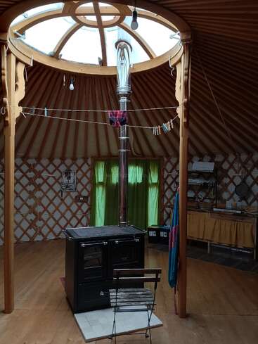 The image depicts the interior of a yurt, featuring a wood-burning stove, a chair, and a clothesline with laundry, set against a backdrop of wooden beams and lattice walls.