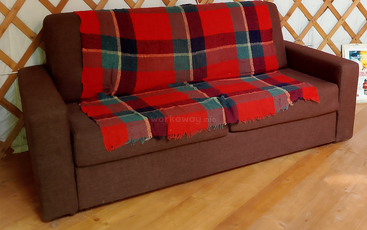 A brown sofa with a colorful red, green, and blue plaid blanket draped over the back and seat, sitting on a wooden floor near a wall.