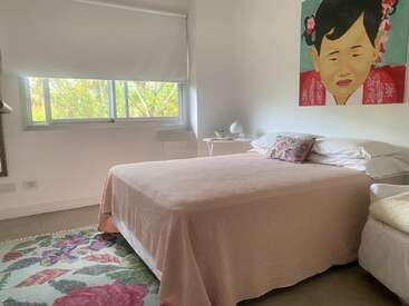 Une chambre douillette avec un lit double recouvert d'une couverture rose pâle, un oreiller à motifs floraux, un tapis coloré, une grande fenêtre, une table de chevet et un portrait d'inspiration asiatique accroché au mur.