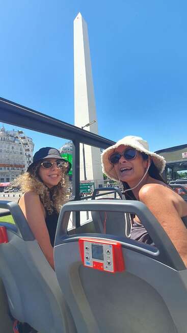 Deux femmes, coiffées de chapeaux et portant des lunettes de soleil, sont assises dans un autocar touristique et sourient. Derrière elles, l'emblématique obélisque de Buenos Aires se dresse fièrement sur fond de ciel bleu limpide.