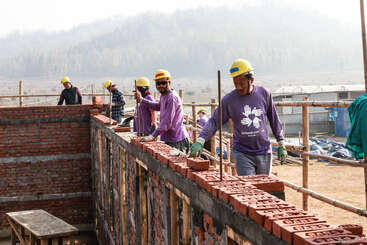 Das Bild zeigt eine Gruppe von Menschen, die auf einer Baustelle arbeiten, mit einem großen Haufen Ziegelsteine und einem Betonfundament im Hintergrund. Die Szene scheint ein im Bau befindliches Gebäude oder eine Struktur zu sein.