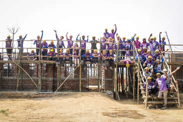 Das Bild zeigt eine Gruppe von Menschen, die ein Bauwerk errichten, wahrscheinlich ein Haus, an dem ein Team von Personen gemeinsam arbeitet. Die Szene spielt sich im Freien ab und ist klar und übersichtlich gestaltet.