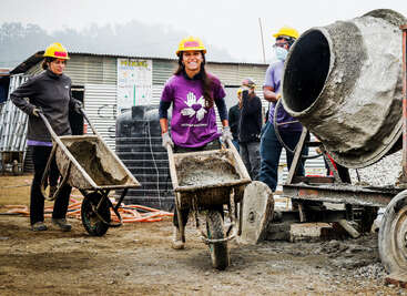 Das Bild zeigt eine Gruppe von Menschen, die auf einer Baustelle arbeiten, Schutzhelme tragen und Schubkarren halten, mit einem großen Zementmischer im Vordergrund.