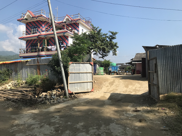 Ein farbenfrohes dreistöckiges Gebäude steht hinter einem Wellblechtor, umgeben von unbefestigten Wegen, verstreuten Steinen, einem schiefen Pfahl, Bäumen und einem klaren blauen Himmel.