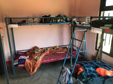 Dieses Bild zeigt einen Schlafsaal mit blauen Metall-Etagenbetten, verstreuten Klamotten, ungemachten Betten, beschrifteten Namensschildern, orangefarbenen Wänden, Sonnenlicht und einer Gitarre am Fenster.