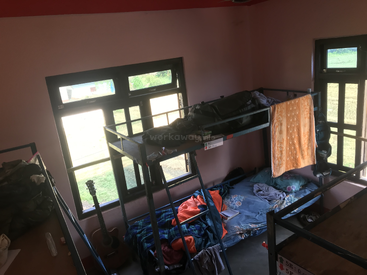 Dieses Bild zeigt einen sonnendurchfluteten Schlafsaal mit rosafarbenen Wänden, Etagenbetten, verstreuten Klamotten, einer Gitarre am Fenster und einem orangefarbenen Handtuch, das über einem Bett hängt.