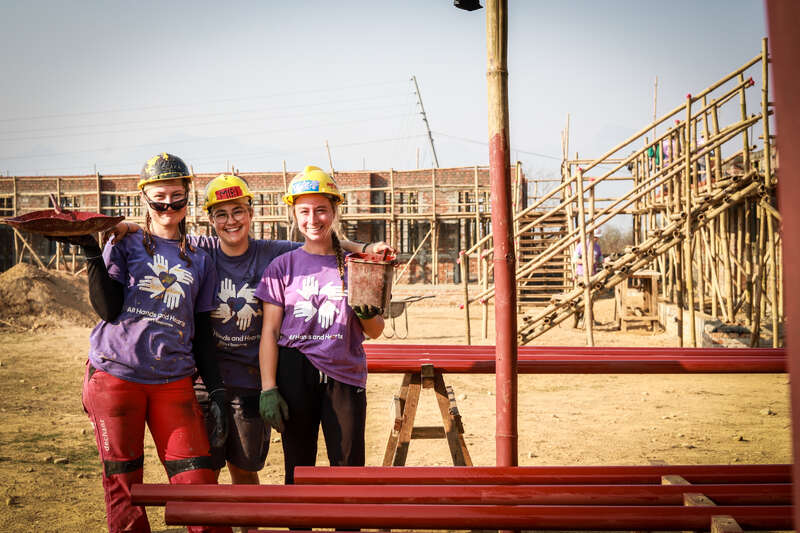 Drei Personen stehen vor einem Gebäude. Zwei von ihnen tragen passende Outfits, die dritte Person trägt ein anderes Outfit und schaut in die Kamera. Das Gebäude ist weiß und hat ein flaches Dach.