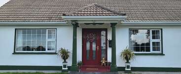 A single-story house with a tiled roof, white exterior, large windows, red door with a wreath, green pillars, potted plants, and a small porch.
