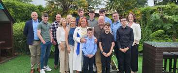 A family group of fifteen stands closely together in a lush green garden, smiling for the camera. Various ages and outfits suggest a special gathering or celebration.