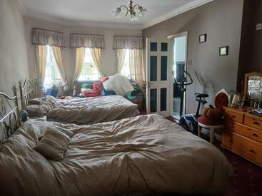 A cozy bedroom features two beds, dresser with mirror, exercise equipment, cluttered corner, three curtained windows, beige walls, and an open door leading to a bathroom.