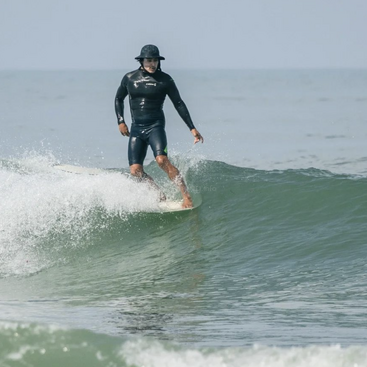 A surfer wearing a black wetsuit and bucket hat confidently rides a small ocean wave. The water is calm, and the sky is clear above.