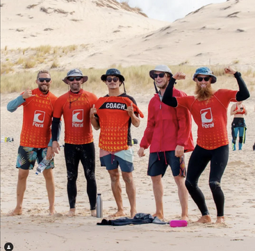 Cinco hombres posan en una playa de arena, vestidos con camisetas y gorras rojas de Feral. Uno de ellos sostiene una camiseta de «Coach». Sonríen, celebrando, rodeados de dunas.