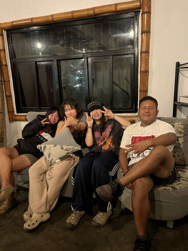 Cuatro personas están sentadas en un sofá dentro de una habitación acogedora, sonriendo y haciendo signos de paz. El ambiente es relajado y alegre, bajo una iluminación cálida.