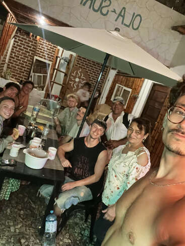 Un alegre grupo de personas se reúne alrededor de una mesa al aire libre por la noche, compartiendo comida y bebida bajo una sombrilla, sonriendo y disfrutando de la compañía mutua.