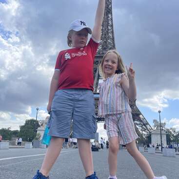 Deux enfants souriants posent devant la Tour Eiffel. Le garçon porte une chemise et un chapeau rouges, la fille porte des rayures. Ils font des signes de paix et profitent de Paris.