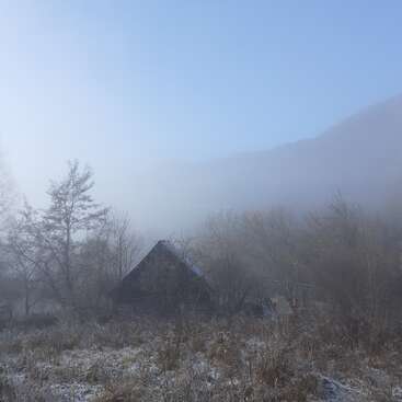 A small wooden house sits quietly among leafless trees on a frosty morning, shrouded in mist, with mountains barely visible under the pale blue sky.