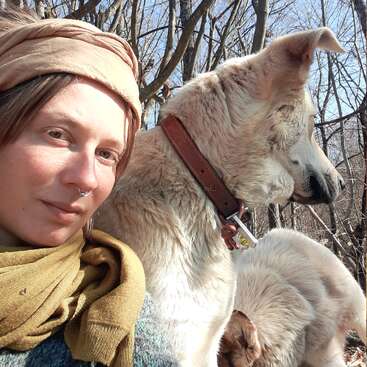 A woman wearing a tan headscarf and green scarf sits outdoors in the sunlight with two cream-colored dogs, surrounded by leafless trees, enjoying nature.