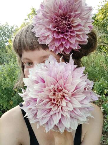 A person poses outdoors, partially hiding their face behind a large, light pink dahlia flower. Another similar flower is tucked in their hair. Lush greenery surrounds them.