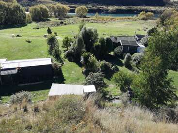 Das Bild zeigt eine ruhige ländliche Landschaft mit einem Haus, einer Scheune und Nebengebäuden, umgeben von üppigem Grün und einem ruhigen Gewässer im Hintergrund.