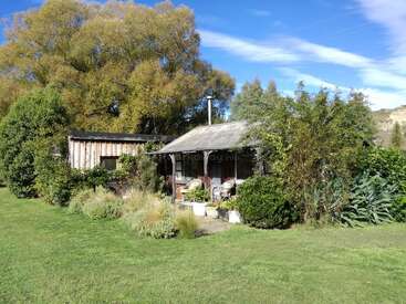 Das Bild zeigt ein kleines, verwittertes Holzhaus mit einer Veranda, umgeben von üppigem Grün und einem strahlend blauen Himmel mit Schäfchenwolken.