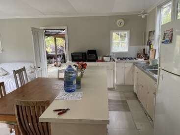 Das Bild zeigt eine Küche mit weißen Schränken und Arbeitsflächen, die mit einem Esstisch, einer Spüle, einem Herd und einem Kühlschrank ausgestattet ist und eine Tür hat, die zu einer Außenterrasse führt.