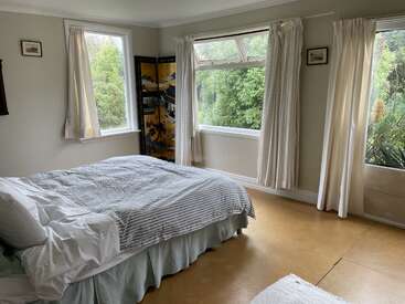 Das Bild zeigt ein ruhiges Schlafzimmer mit einem Bett, Fenstern und Vorhängen, mit einer weißen Bettdecke und einem gelben Paravent in der Ecke, umgeben von üppigem Grün draußen.