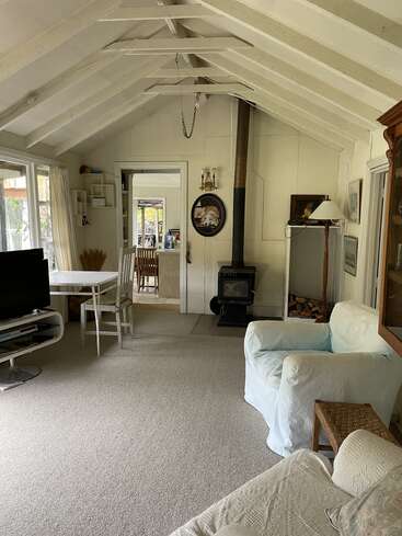 Das Bild zeigt ein gemütliches Wohnzimmer mit weißen Wänden, einer gewölbten Decke und einem Holzofen, mit einer Couch, einem Tisch, einem Fernseher und einer Tür zur Küche.