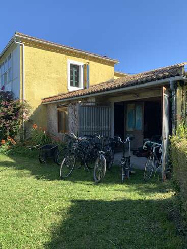 L'image représente une maison jaune à deux étages avec un toit en tuiles, dotée d'un patio couvert et d'une rangée de bicyclettes garées devant.