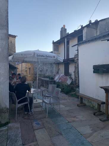 L'image représente un espace extérieur serein dans un village pittoresque, avec une table et des chaises, un parasol et un banc, entouré de bâtiments rustiques et d'un ciel bleu clair.