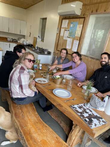 Five people are seated around a rustic wooden table in a cozy kitchen, sharing a meal and smiling. A dog sits nearby, adding warmth to the scene.