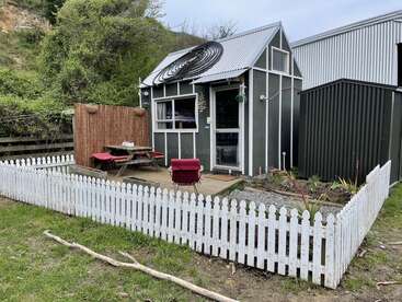 A small, cozy tiny house with a metal roof, surrounded by a white picket fence. Outdoor patio with picnic table, chair, and garden area. Peaceful setting.