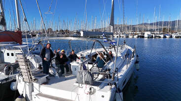 Un groupe de personnes se détendent et sourient sur un voilier amarré dans une marina, entouré de nombreux autres bateaux, par une journée claire et ensoleillée.