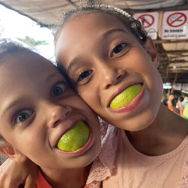 Duas crianças sorridentes posam juntas, cada uma com um limão verde na boca, brincando e imitando grandes sorrisos. O plano de fundo mostra uma área coberta com sinalização.