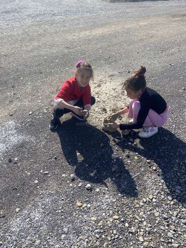Duas meninas estão brincando juntas em um terreno rochoso e ensolarado. Elas estão agachadas, sorrindo e construindo algo com terra, curtindo a atividade ao ar livre.