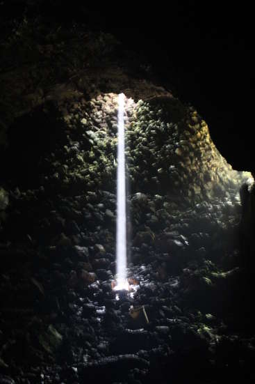 The image depicts a cave with a vertical beam of light shining down from the top, illuminating rocks and moss, creating a striking contrast between light and darkness.