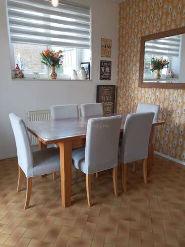A imagem mostra uma sala de jantar com uma mesa e cadeiras de madeira, uma janela e um espelho na parede, com uma atmosfera calorosa e convidativa.