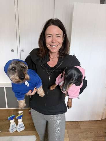 A smiling woman in a black jacket holds two small dogs. One wears a blue hoodie, the other pink. Cleaning supplies are visible on the wooden floor.