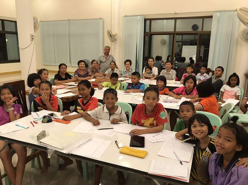 The image depicts a classroom with approximately 20 children and several adults seated at tables, engaged in a learning activity, with a calm and studious atmosphere.