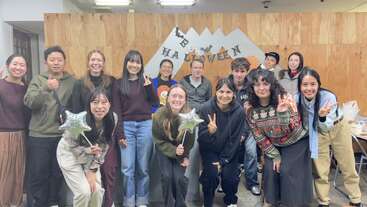 Un alegre grupo de personas posa en el interior, sonriendo y haciendo el signo de la paz. Detrás de ellos, una decoración con motivos de Halloween cuelga de una pared de madera. Dos de ellos sostienen globos con forma de estrella.