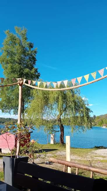 Bandeiras triangulares coloridas estão penduradas acima de um lago tranquilo, cercado por árvores verdes exuberantes, sob um céu azul claro, com um prédio vermelho e uma paisagem pacífica.