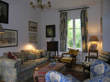 Esta aconchegante sala de estar apresenta sofás estampados, cadeiras antigas, uma mesa de centro de madeira, arte na parede, um lustre e uma janela iluminada pelo sol com cortinas azuis e amarelas.