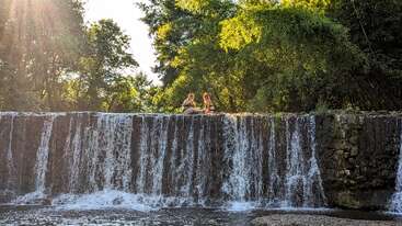 Zwei Menschen sitzen auf einem kleinen Wasserfall, umgeben von üppig grünen Bäumen. Das Sonnenlicht fällt durch das Laub und schafft eine heitere und sommerliche Atmosphäre in der Natur.