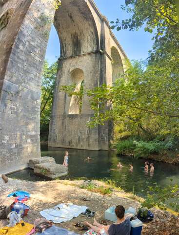 Die Menschen schwimmen und entspannen sich unter einem großen steinernen Viadukt, umgeben von üppigem Grün. Handtücher und Habseligkeiten liegen auf dem Boden verstreut und erinnern an eine friedliche Sommerszene.