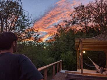 Eine Person bewundert einen atemberaubenden Sonnenuntergang mit leuchtend orangefarbenen Wolken und blauem Himmel, umgeben von Bäumen, einem Holzdeck und einer gemütlichen Hängematte unter einem Unterstand.