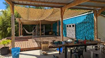 Ein Holzdeck mit Hängematten und einem Tarnnetzdach steht neben einem blauen Wellblechgebäude. In der Nähe befinden sich Arbeitsgeräte und Stühle in einem geschützten Arbeitsbereich.